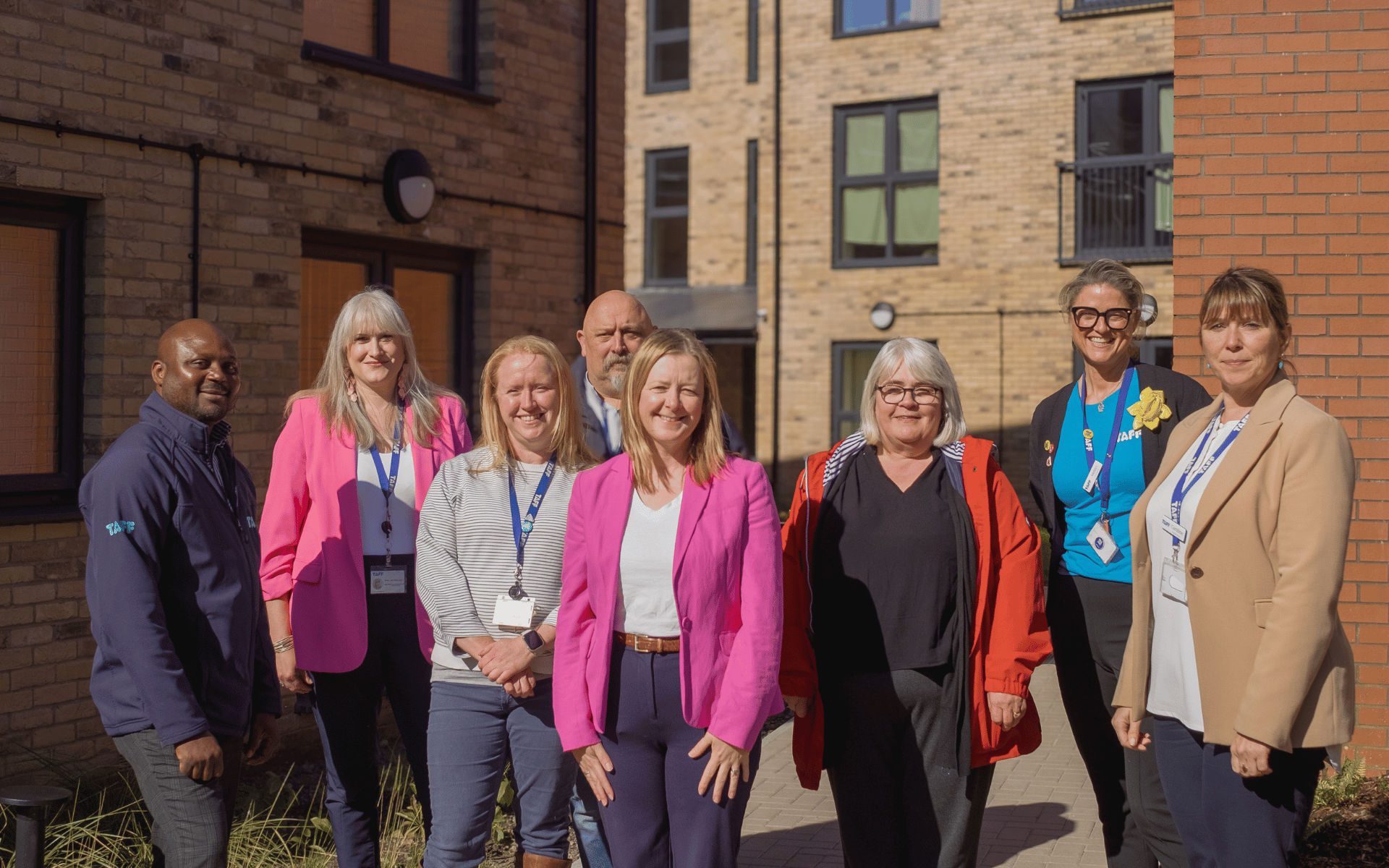 A group of eight adults smiling and posing for a photo outdoors in a courtyard between modern brick residential buildings.