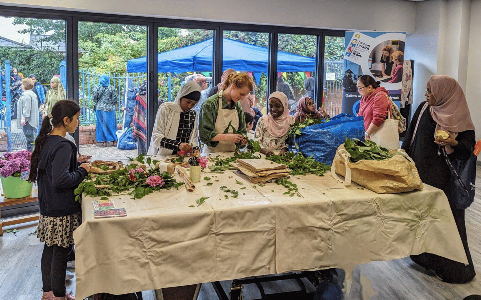 A diverse group of women and children gathered around a large table indoors, participating in a flower and greenery crafting activity. Through large glass doors behind them, a busy outdoor community event with blue canopy tents is visible.