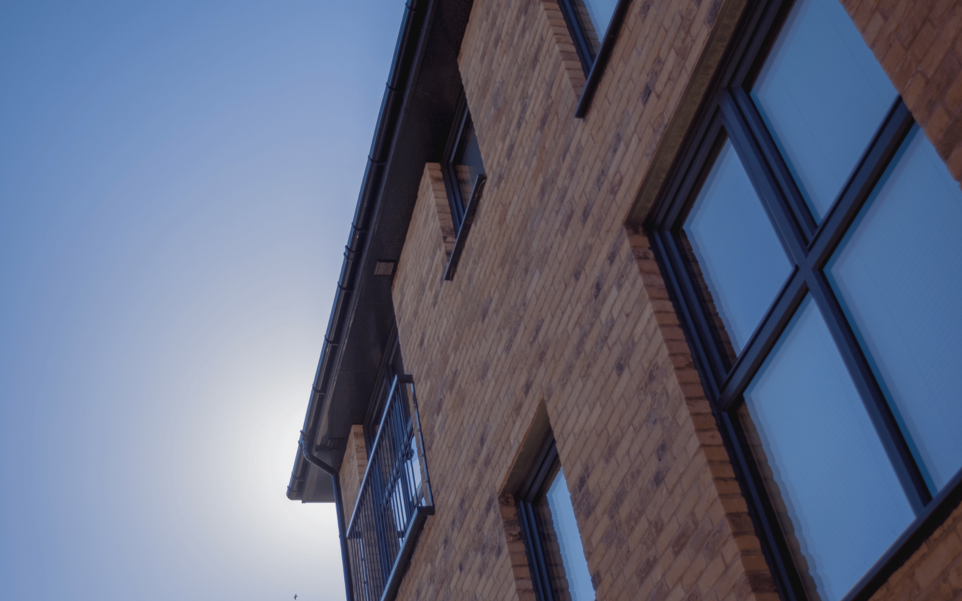 A low-angle view looking up at the exterior of a modern brown brick building with large, dark-framed windows against a clear blue sky.
