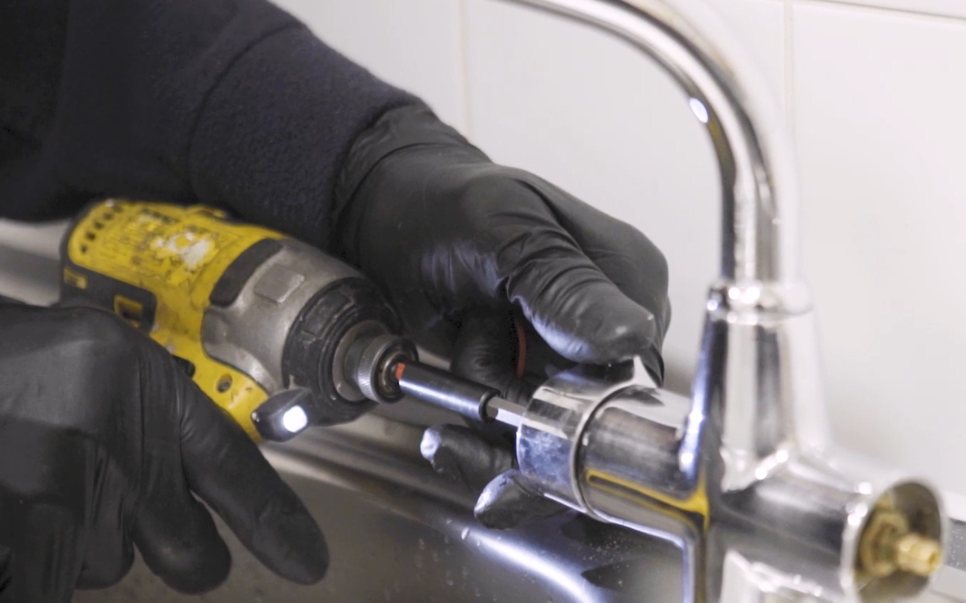 A close-up of gloved hands using a yellow power drill to repair or install a chrome kitchen tap.