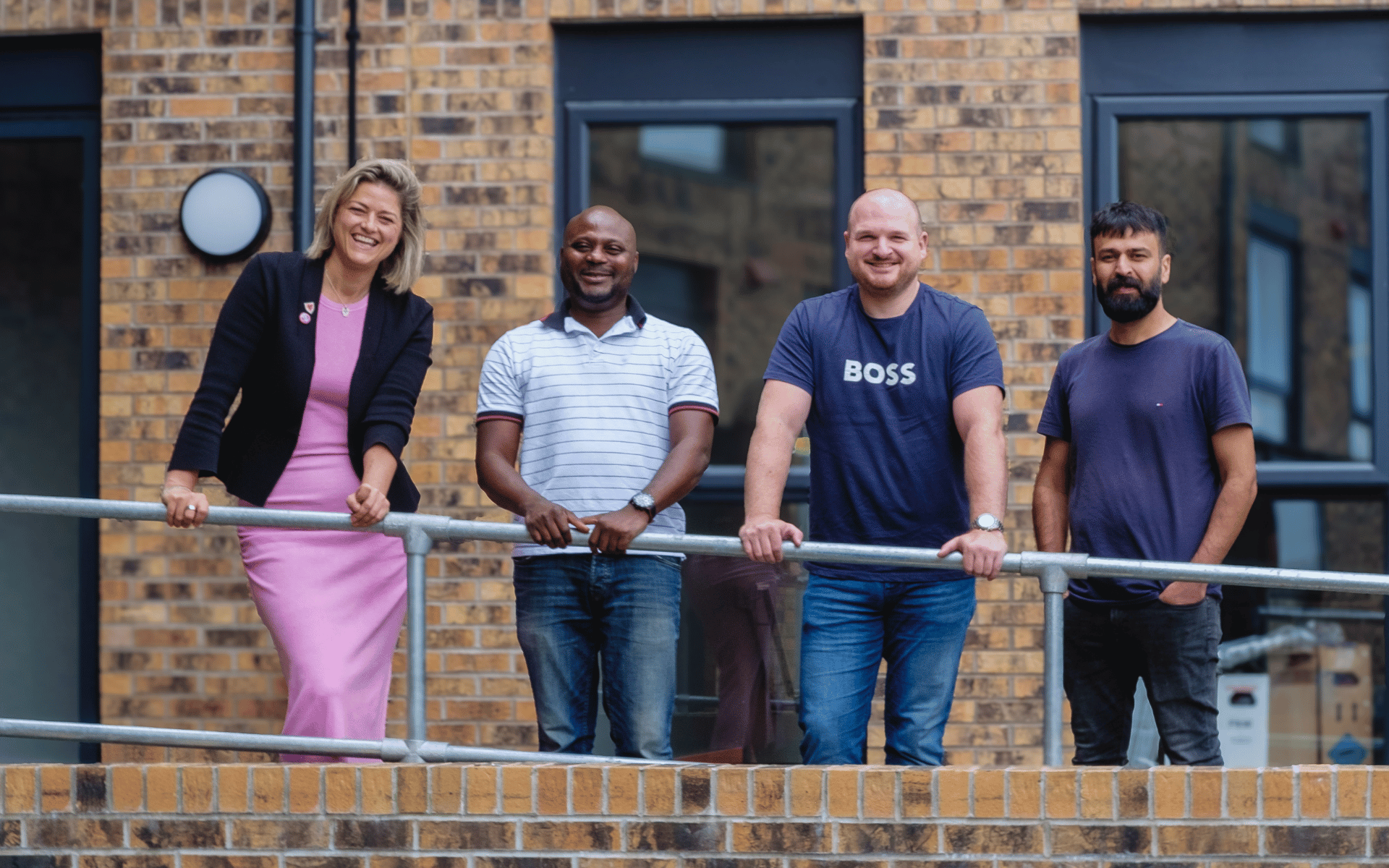 A group of four adults smiling and posing together behind a metal railing outside a modern brick building.