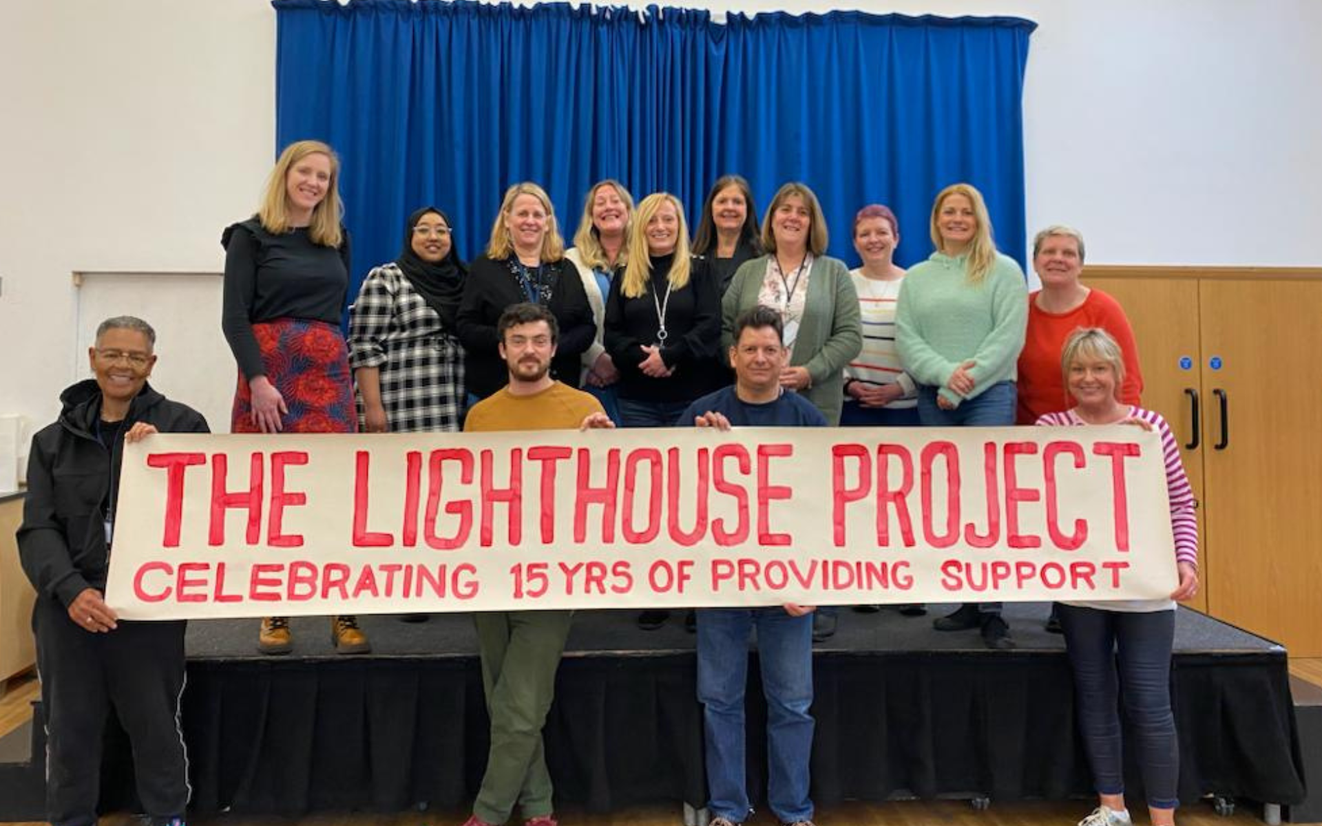 A group of people posing on and in front of a stage holding a large banner that reads "THE LIGHTHOUSE PROJECT CELEBRATING 15 YRS OF PROVIDING SUPPORT".