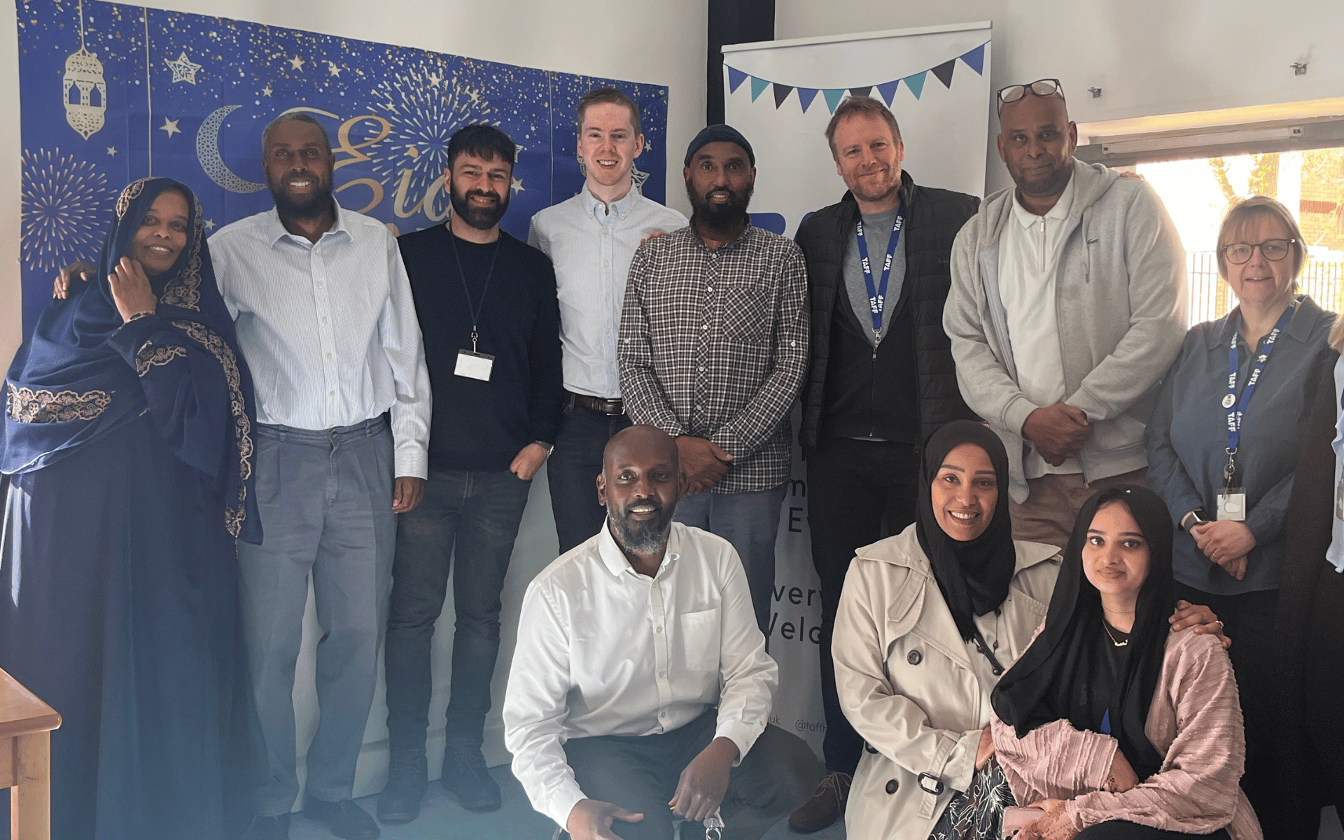 A diverse group of eleven adults smiling together for a group portrait indoors, posing in front of a blue and gold banner decorating the room for an Eid celebration.