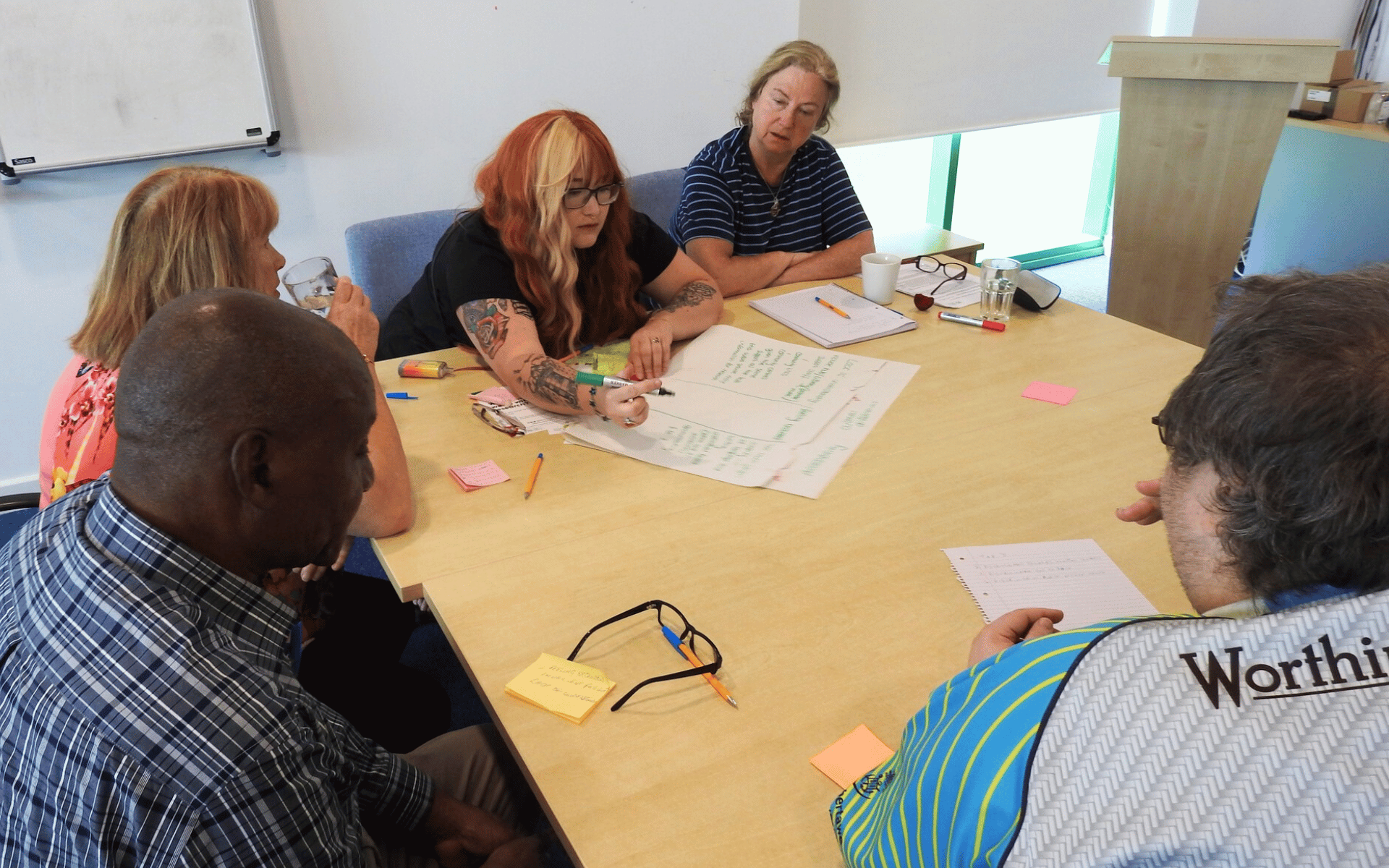 A group of five adults sitting around a table in a bright room, actively engaged in a meeting or workshop as they look at a large sheet of paper with handwritten notes.