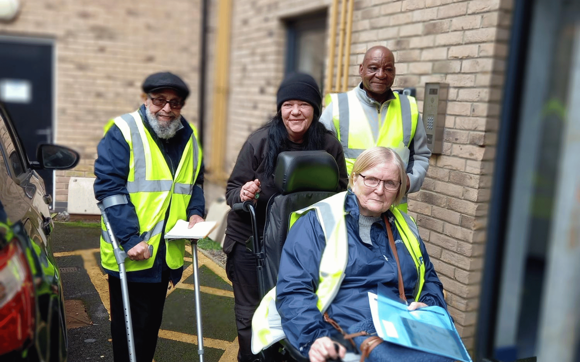 A group of four adults, three of whom are wearing yellow high-visibility vests, smiling for a photo outdoors next to a brick building. One person is using a crutch and another is seated in a wheelchair.