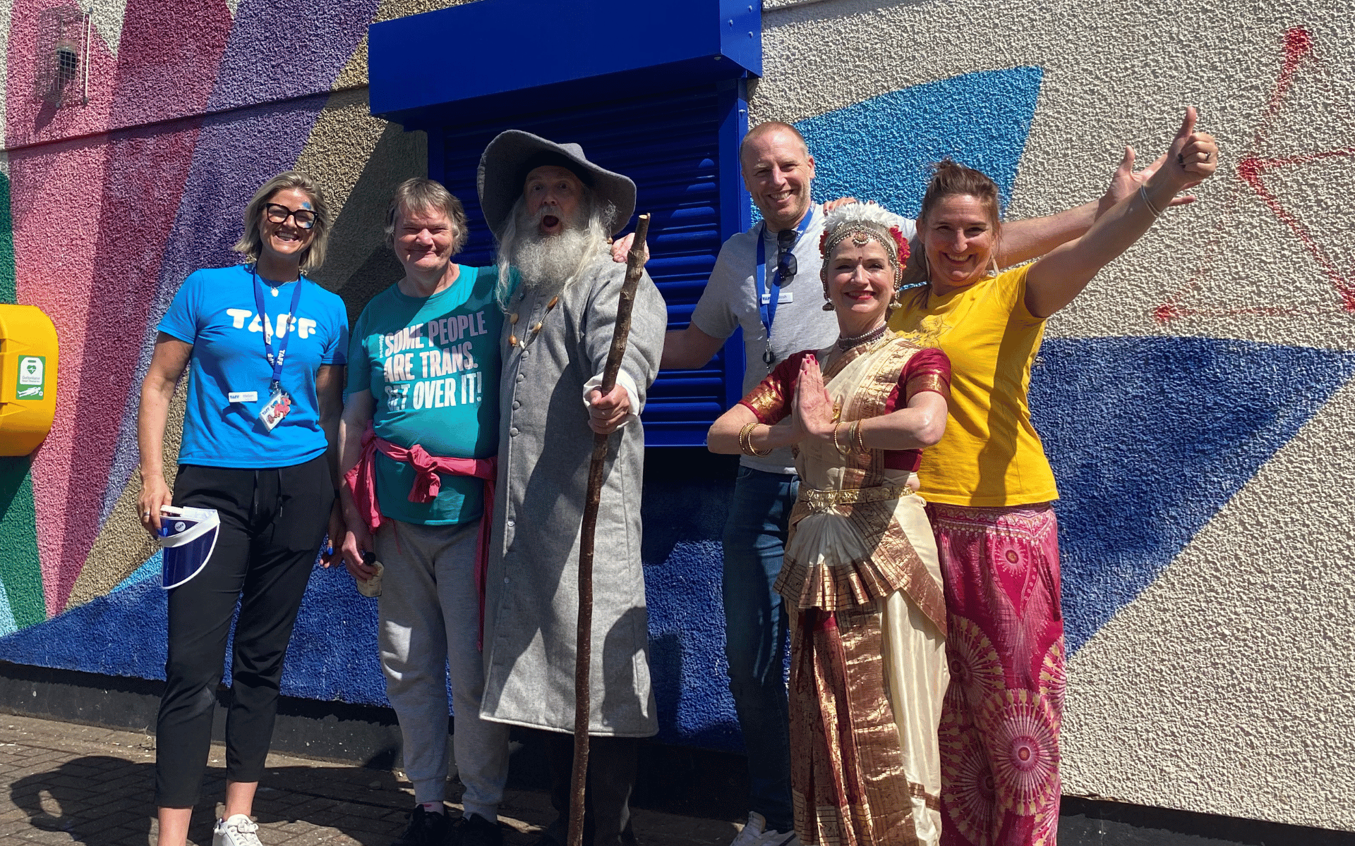 A diverse group of six adults, including some in costumes like a wizard and traditional Indian attire, posing cheerfully together outdoors in front of a brightly painted geometric mural.
