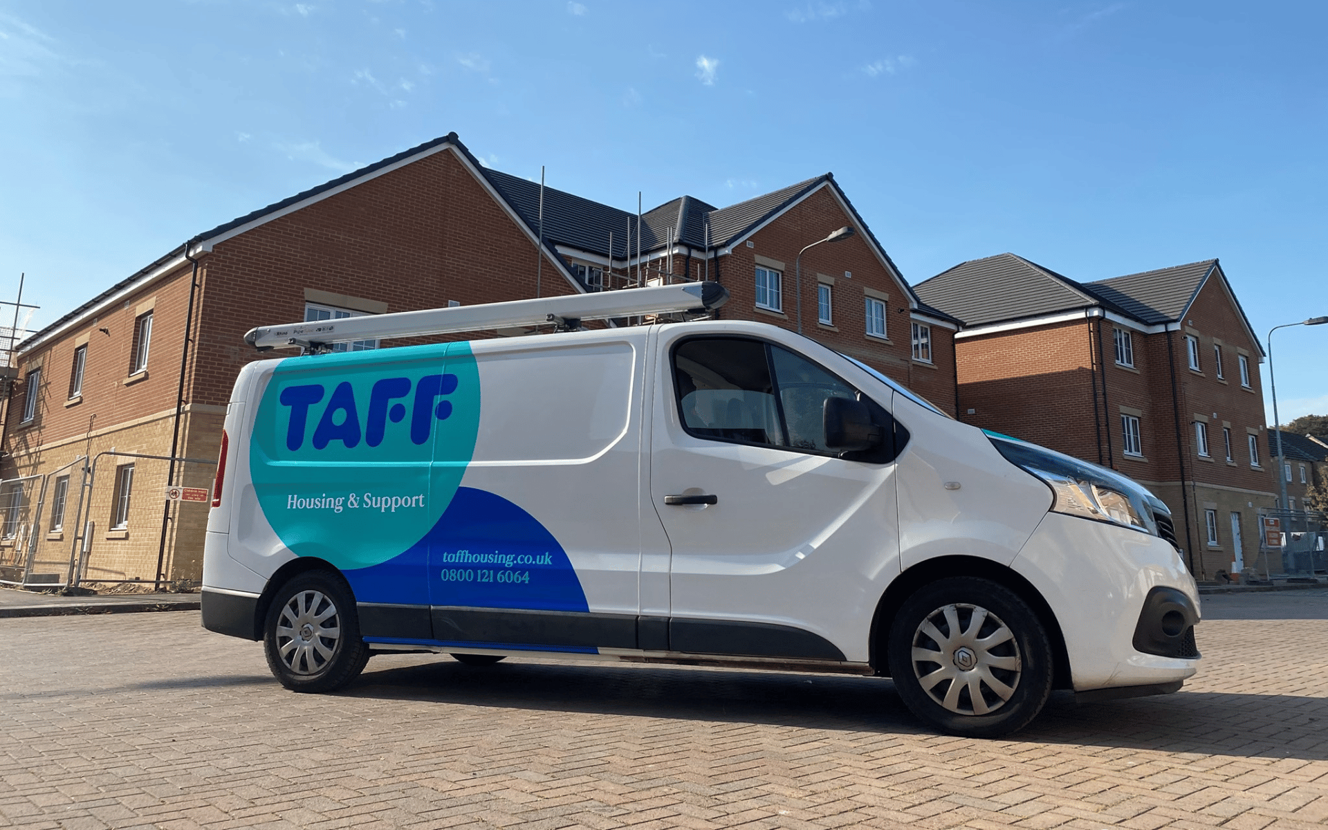 A white TAFF Housing & Support van parked on a block-paved area in front of modern brick residential buildings under a clear blue sky.