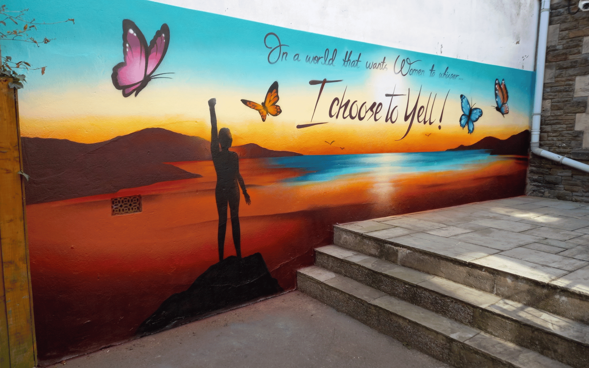 A vibrant outdoor mural depicting a silhouette of a person with a raised fist against a colorful sunset over water. The mural features flying butterflies and the quote, "In a world that wants Women to whisper... I choose to Yell!".